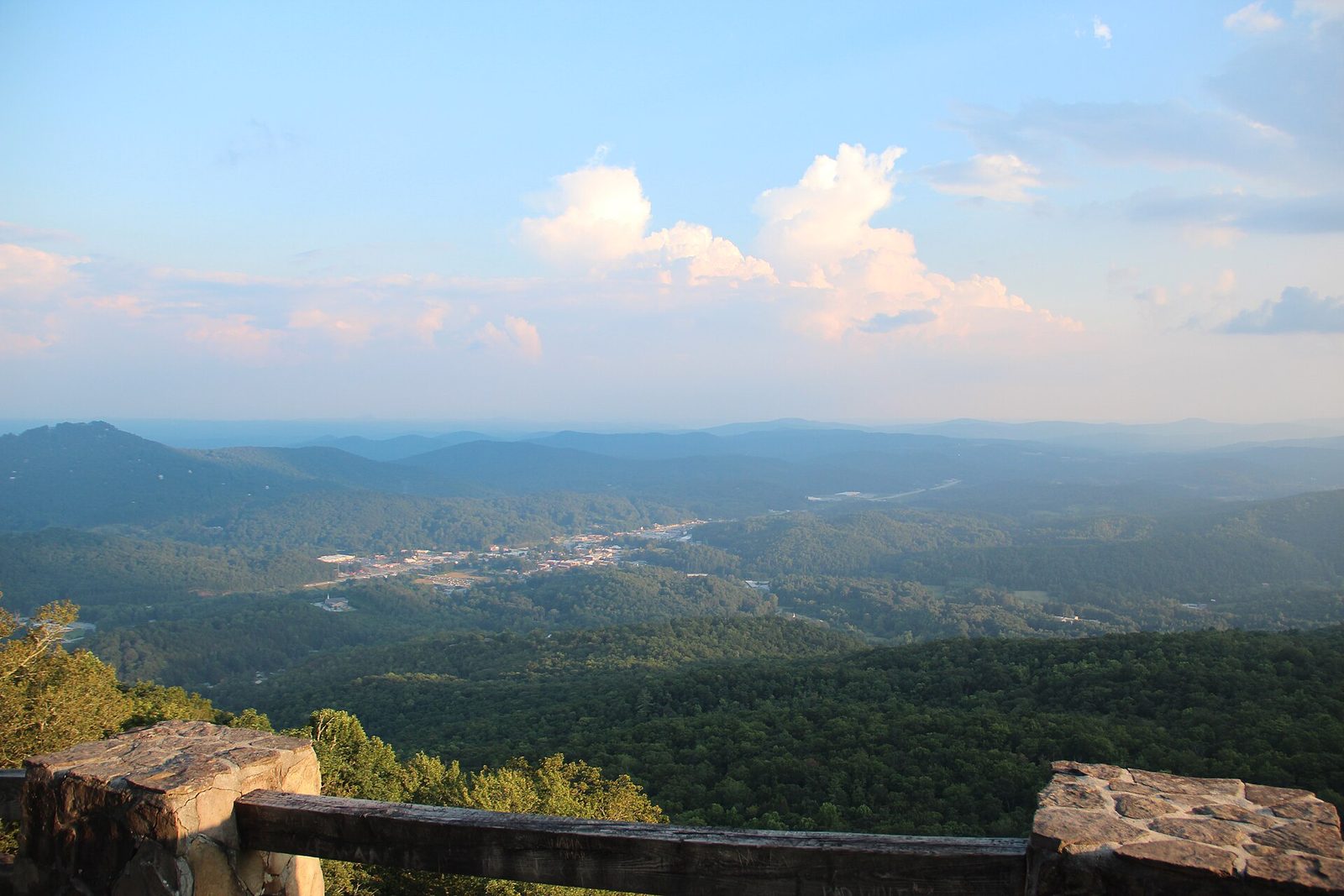 A long view over forested blue ridges and the town of Clayton in the valley below, seen over a stone and timber overlook wall at Black Rock Mountain State Park