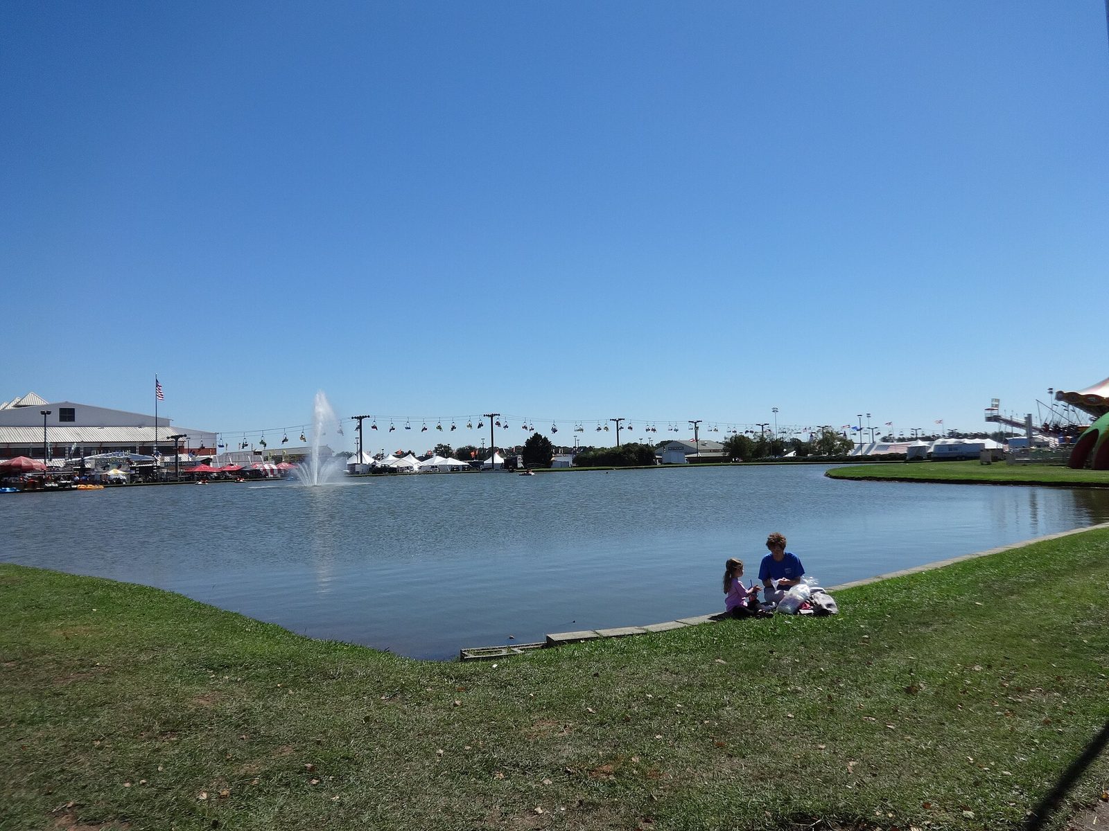 The lake at the Georgia National Fairgrounds in Perry with a fountain, sky ride cabins strung across behind it, exhibit halls and tents in the distance under a clear blue sky