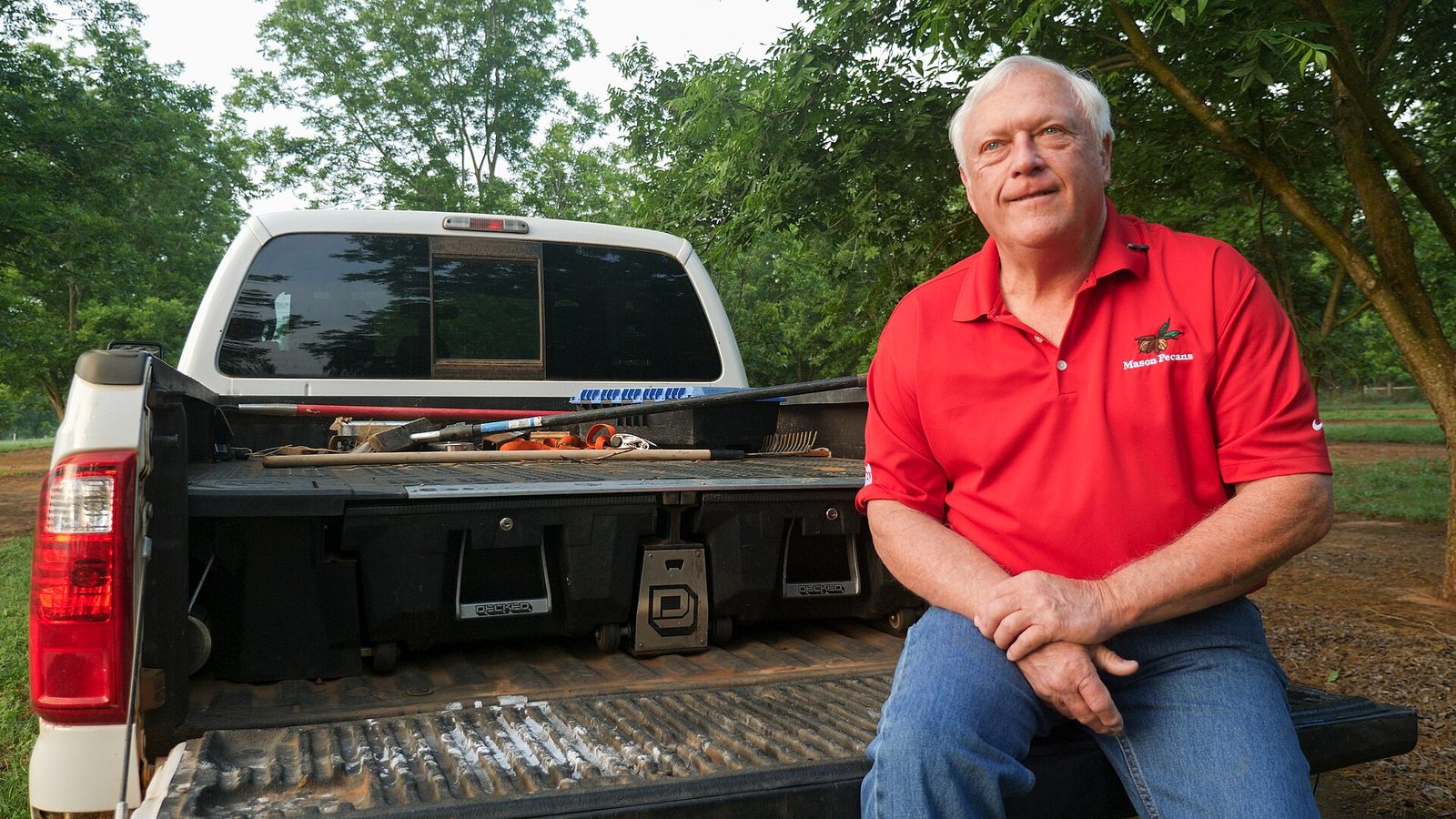 A pecan grower in a red Mason Pecans polo shirt sitting on the tailgate of a white pickup truck in his orchard in south Georgia