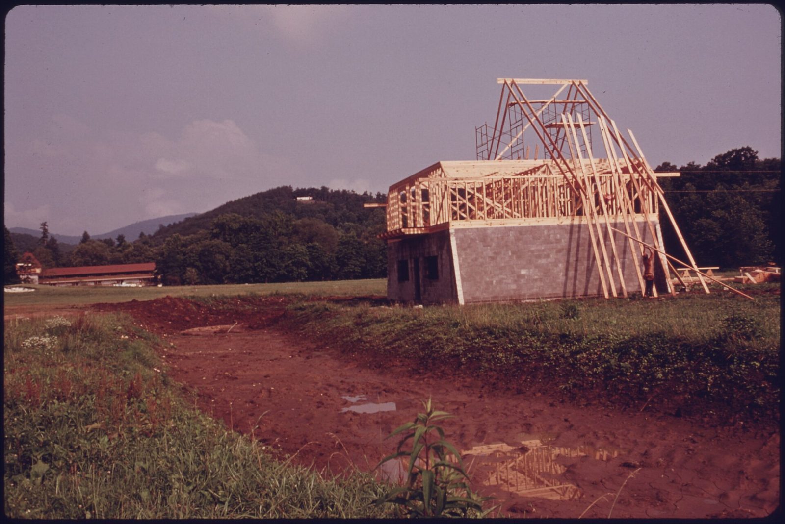 A timber-framed alpine-style building under construction in a field of red clay outside Helen, Georgia, with wooded mountains behind, photographed in the early 1970s