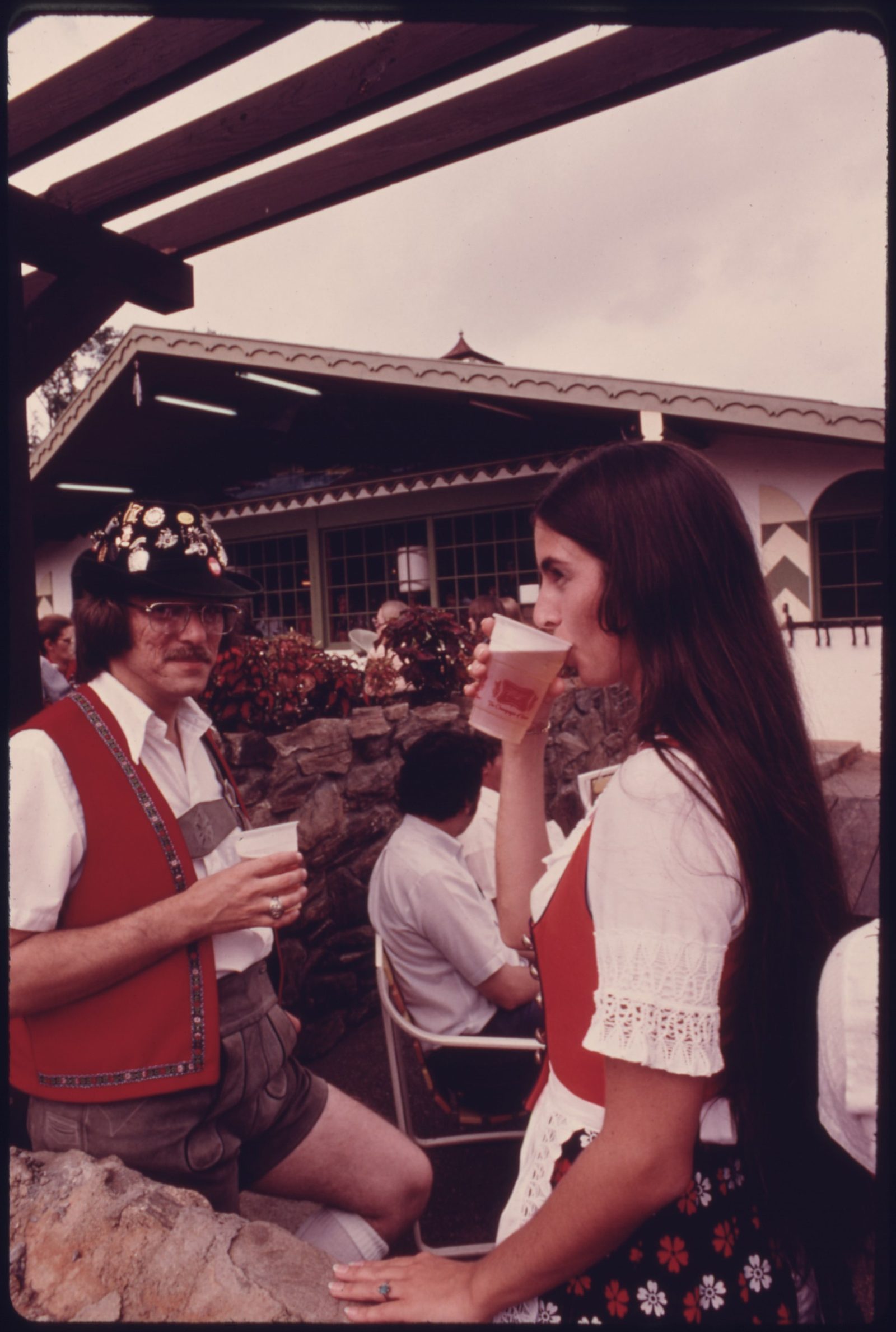 Two people in Bavarian dress, a man in a red vest and lederhosen and a woman in a red dirndl bodice, drinking from cups outside an alpine-style building at Helen's Oktoberfest in the 1970s