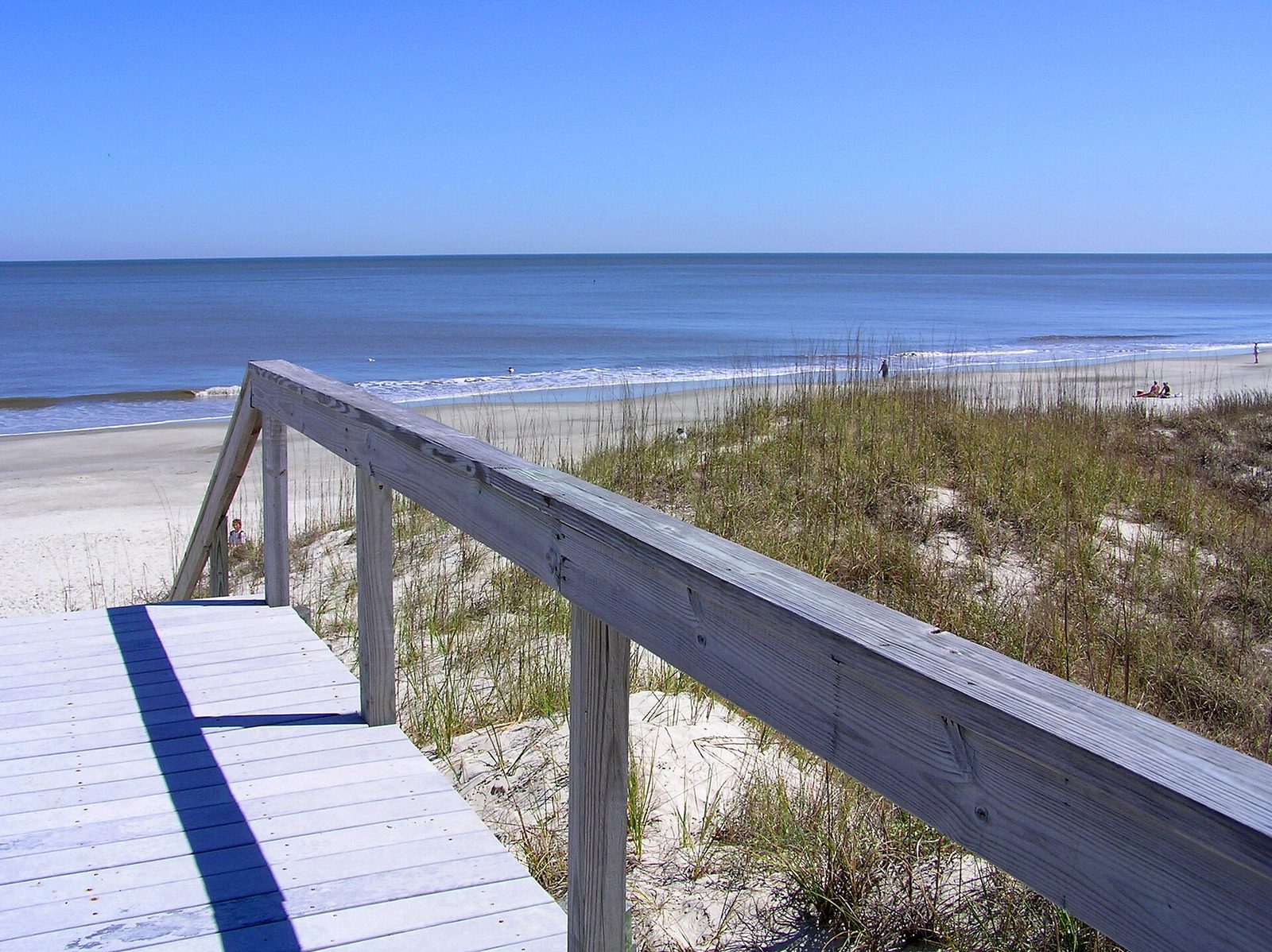A weathered wooden boardwalk over sea oats leading onto a wide, almost empty beach on Jekyll Island under a clear blue sky