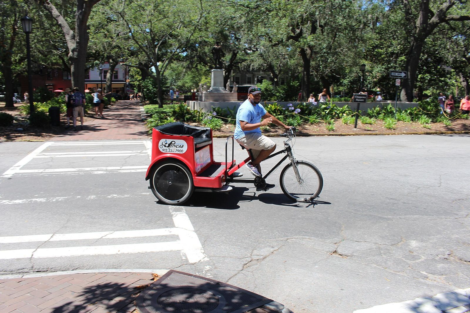 A red pedicab crossing an intersection beside Chippewa Square in Savannah, with live oaks hung with Spanish moss and a monument behind