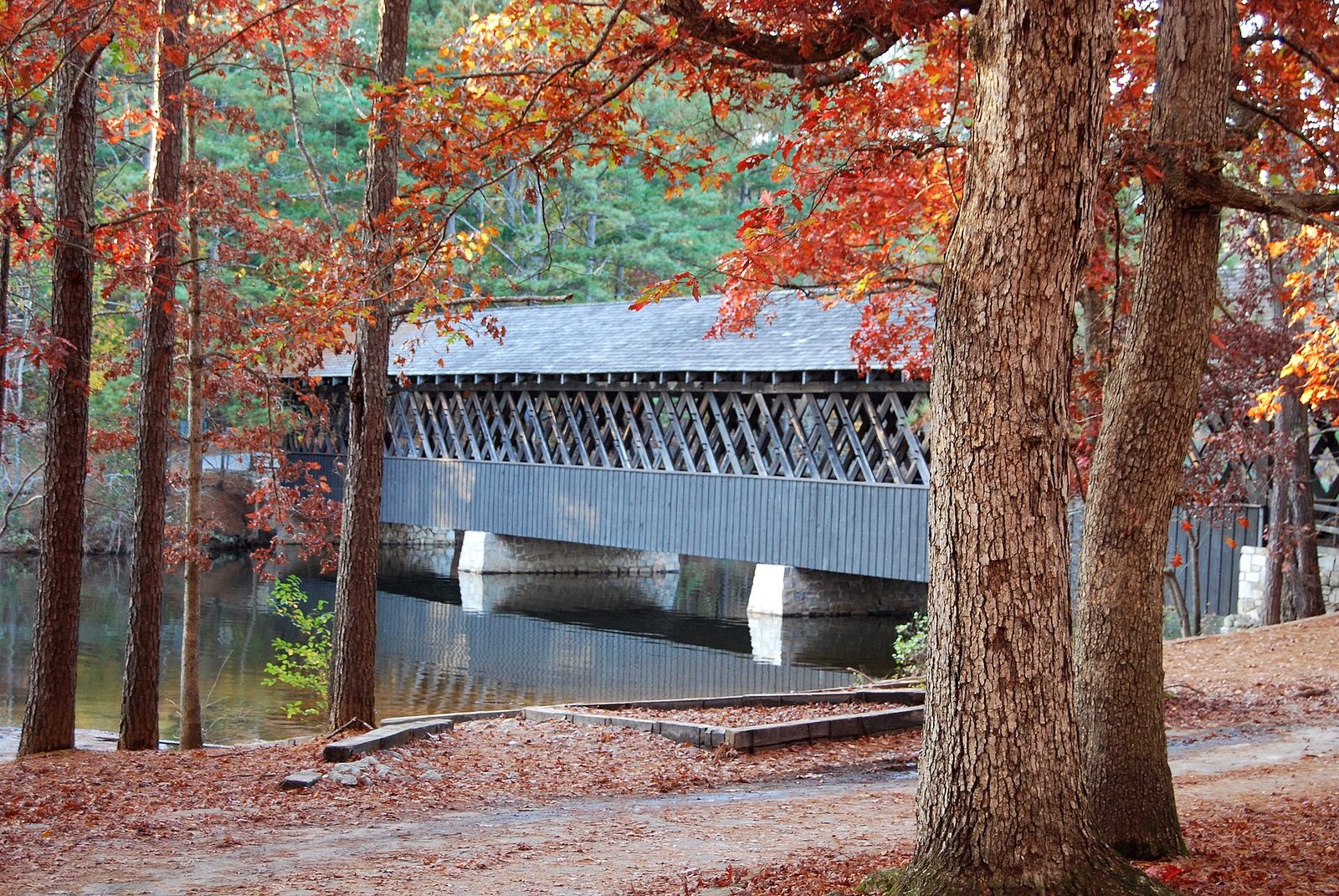 A wooden covered bridge crossing still water at Stone Mountain Park, framed by pines and oaks in red autumn color